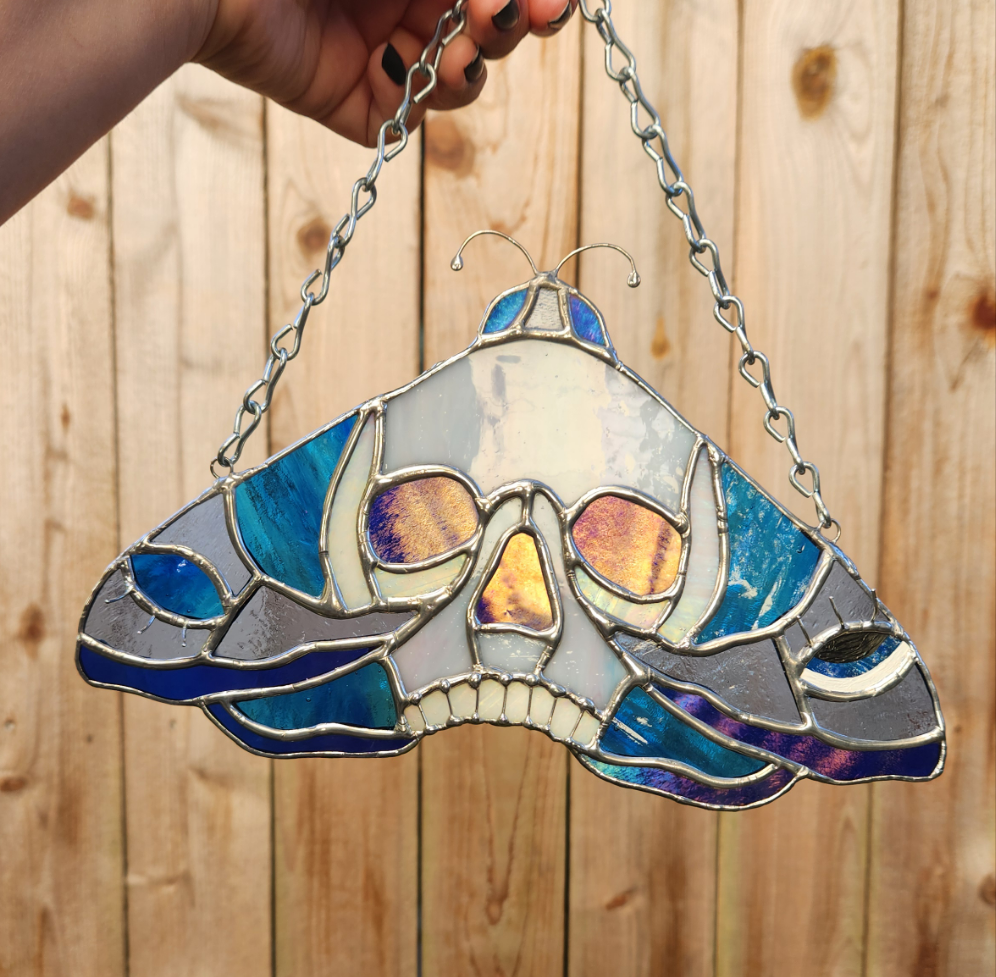 Stained Glass Skull Moth Suncatcher held in front of a wooden fence. The moth is silver with blue toned glass.