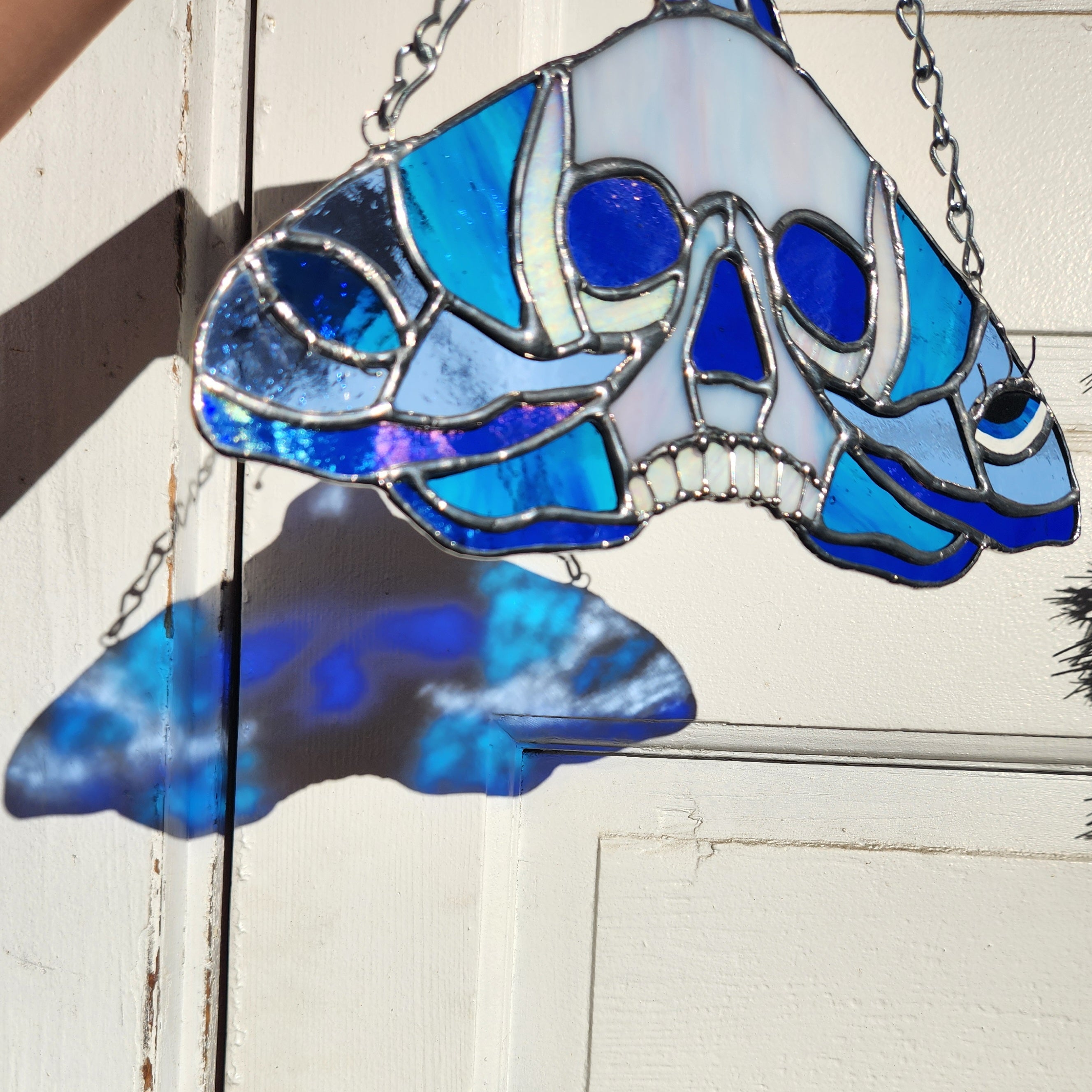 Stained Glass Skull Moth Suncatcher held in front of a door with sunlight shining through. The moth is silver with blue toned glass.