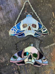 Stained Glass Skull Moth Suncatchers on table. The top moth is silver and blue toned glass, the bottom is pink and purple glass with black patina 