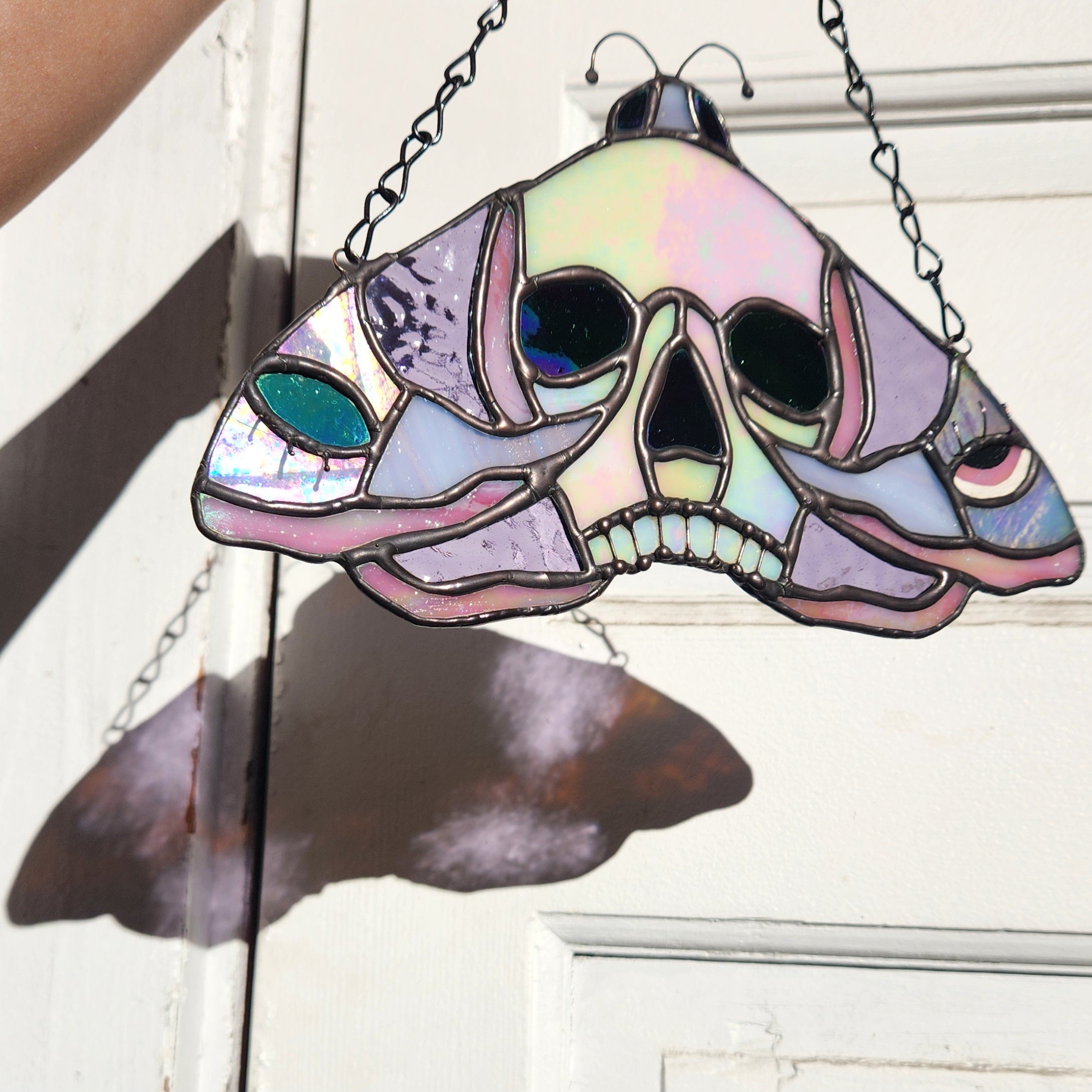 Stained Glass Skull Moth Suncatcher held in front of a door with sunlight shining through. The moth is pink and purple glass with black patina.