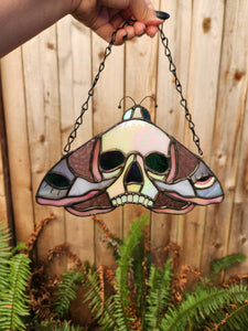 Stained Glass Skull Moth Suncatcher held in front of a wooden fence. The moth is pink and purple glass with black patina.