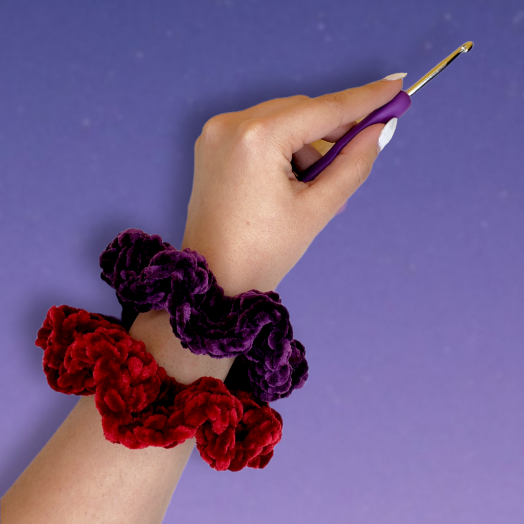 Deep red (wine) and dark purple (royal) crocheted velvet scrunchies on wrist. Hand holding crochet hook.