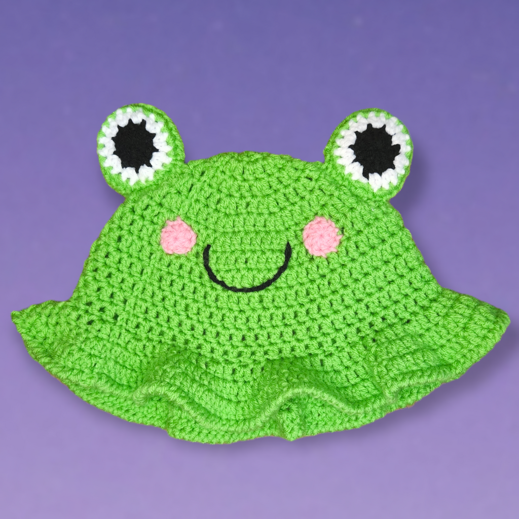 Front view of the Hippity Hop— Frog Hat. The main color is lime green, it has black and white eyes that stick out of the top. Two light pink cheeks and a black smile are on the hat's body. It has a wavy brim.